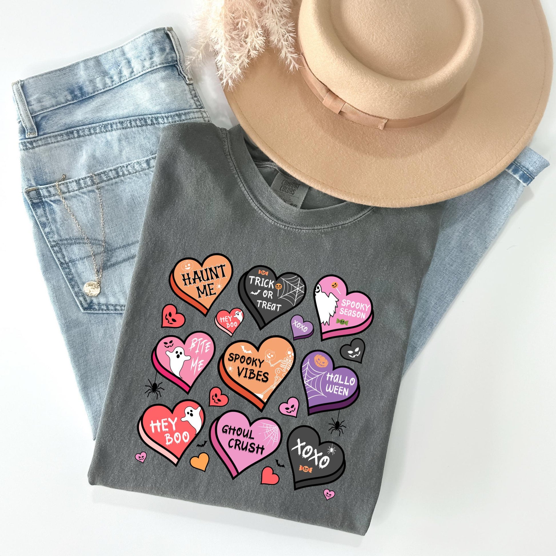 a gray t-shirt with a variety of Halloween-themed heart shapes and text, including phrases like "HAINT ME", "TRICK OR TREAT", "SPOOKY VIBES", "HEY BOO", "CHULO CRUSH", "XOXO", and "HALLO WED". The t-shirt is placed on top of a pair of blue jeans and a beige hat, creating a casual and festive Halloween-inspired outfit.