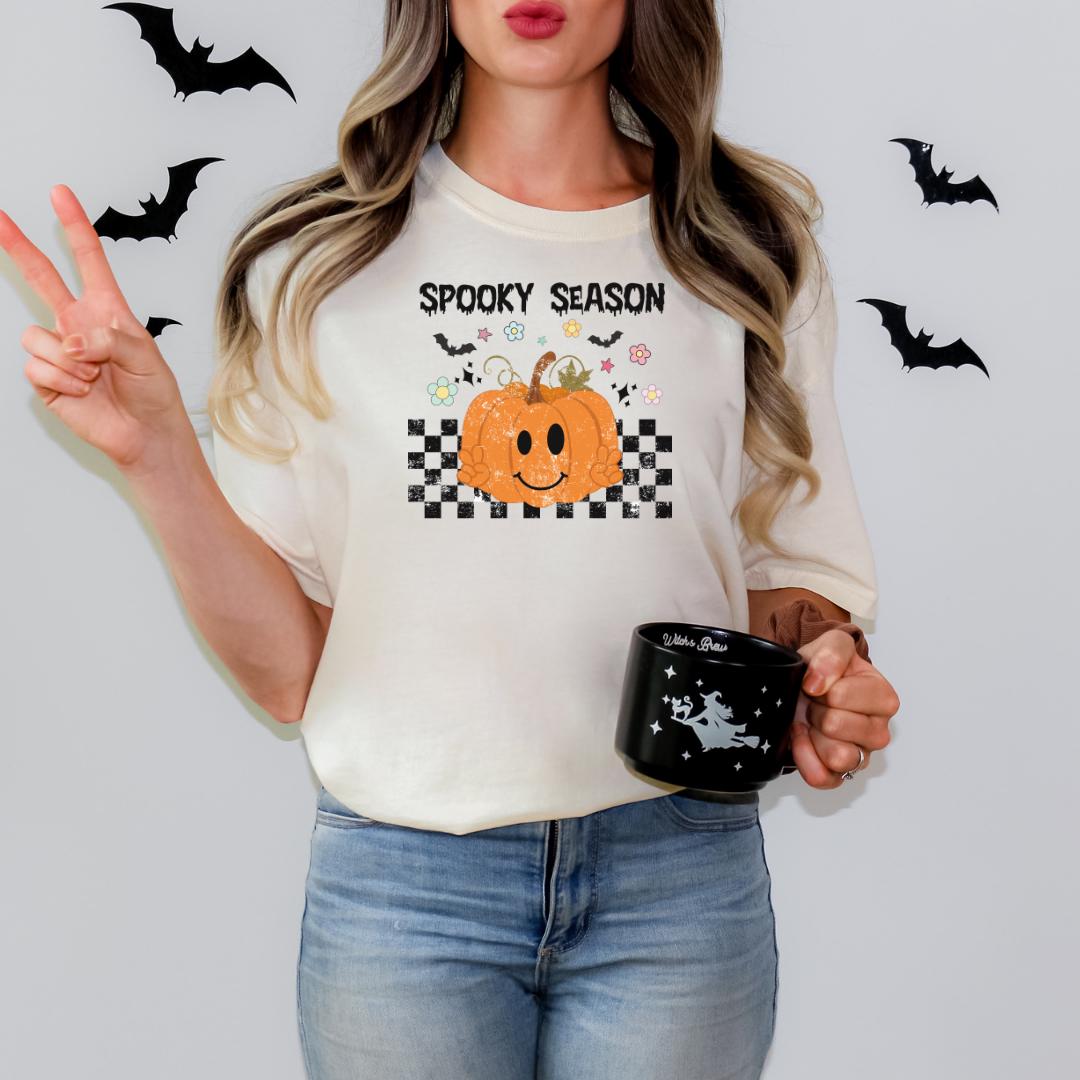 A woman wearing a white t-shirt with a pumpkin graphic and the text "Spooky Season" is holding a black mug with a Halloween-themed design.