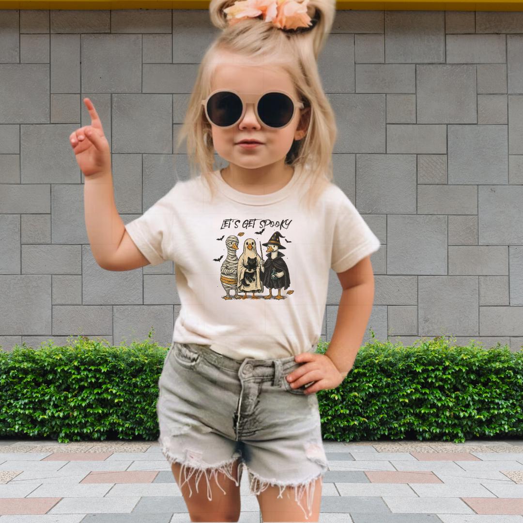A young girl wearing a white t-shirt with a graphic design, sunglasses, and denim shorts is standing in front of a brick wall.