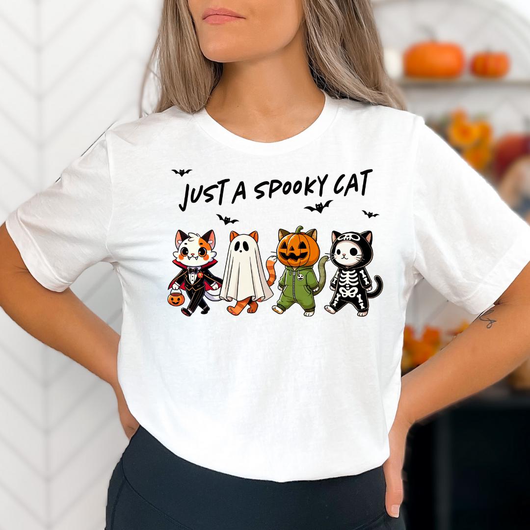 A woman wearing a white t-shirt with a graphic design featuring four cartoon cats dressed in Halloween costumes, including a skeleton, a ghost, a jack-o-lantern, and a witch.