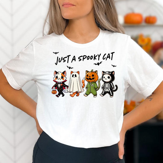 A woman wearing a white t-shirt with a graphic design featuring four cartoon cats dressed in Halloween costumes, including a skeleton, a ghost, a jack-o-lantern, and a witch.