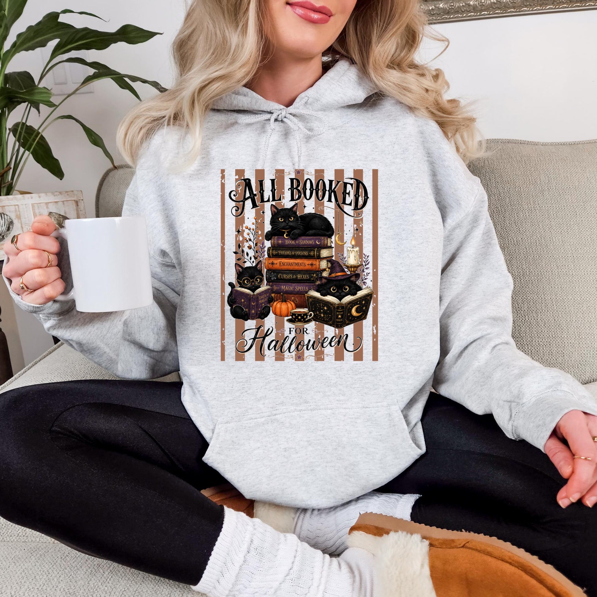 A woman wearing a grey hoodie with a Halloween-themed graphic and holding a mug is sitting on a couch.