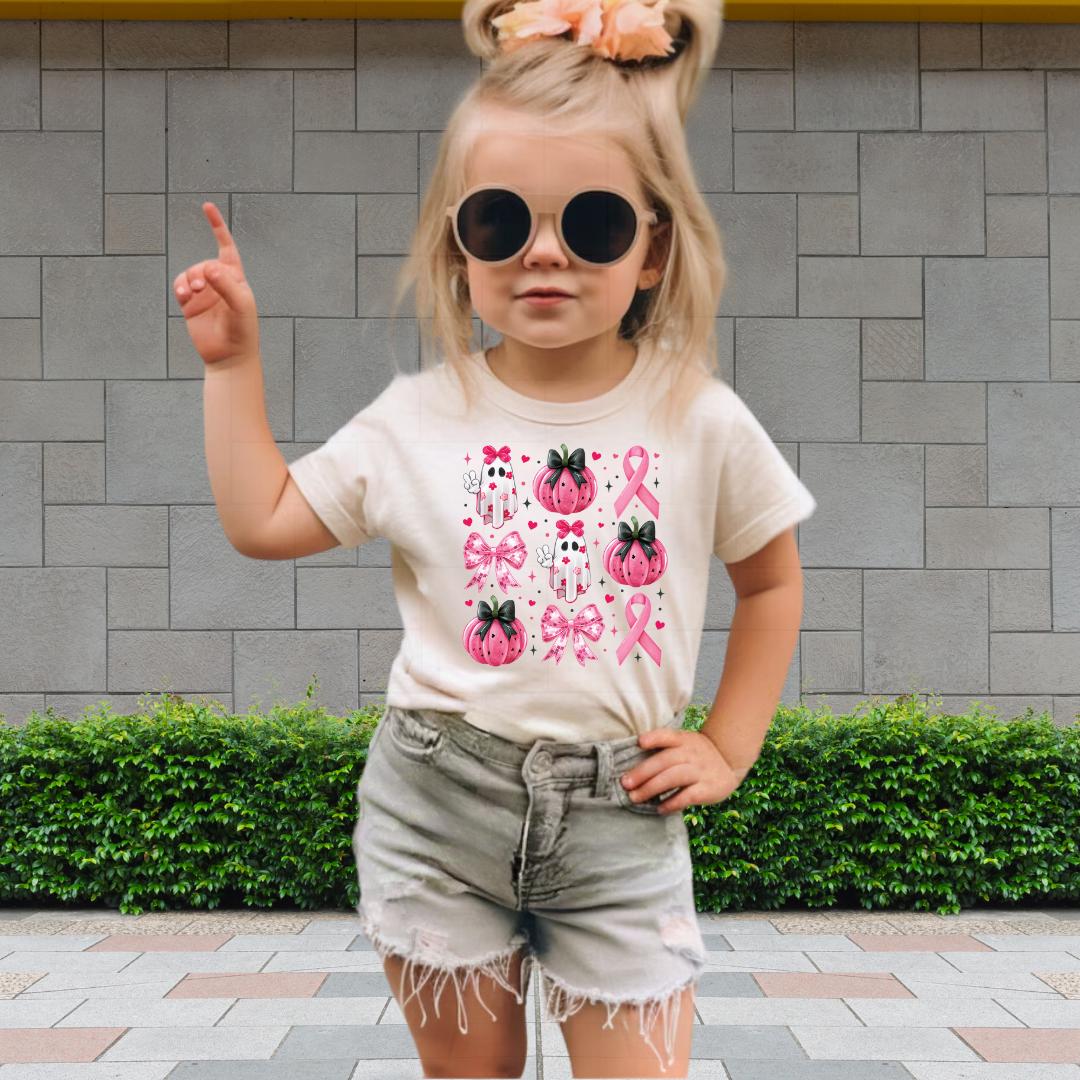 A young girl wearing a white t-shirt with pink pumpkins and a pink ribbon design, sunglasses, and denim shorts is standing in front of a gray brick wall.