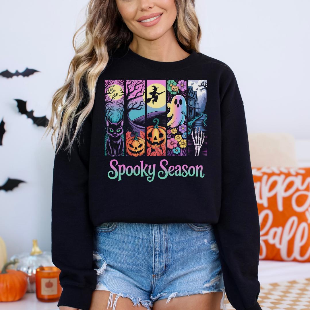 A woman wearing a black sweatshirt with a graphic design featuring Halloween-themed illustrations, including bats, pumpkins, a skeleton, and a witch on a broomstick.