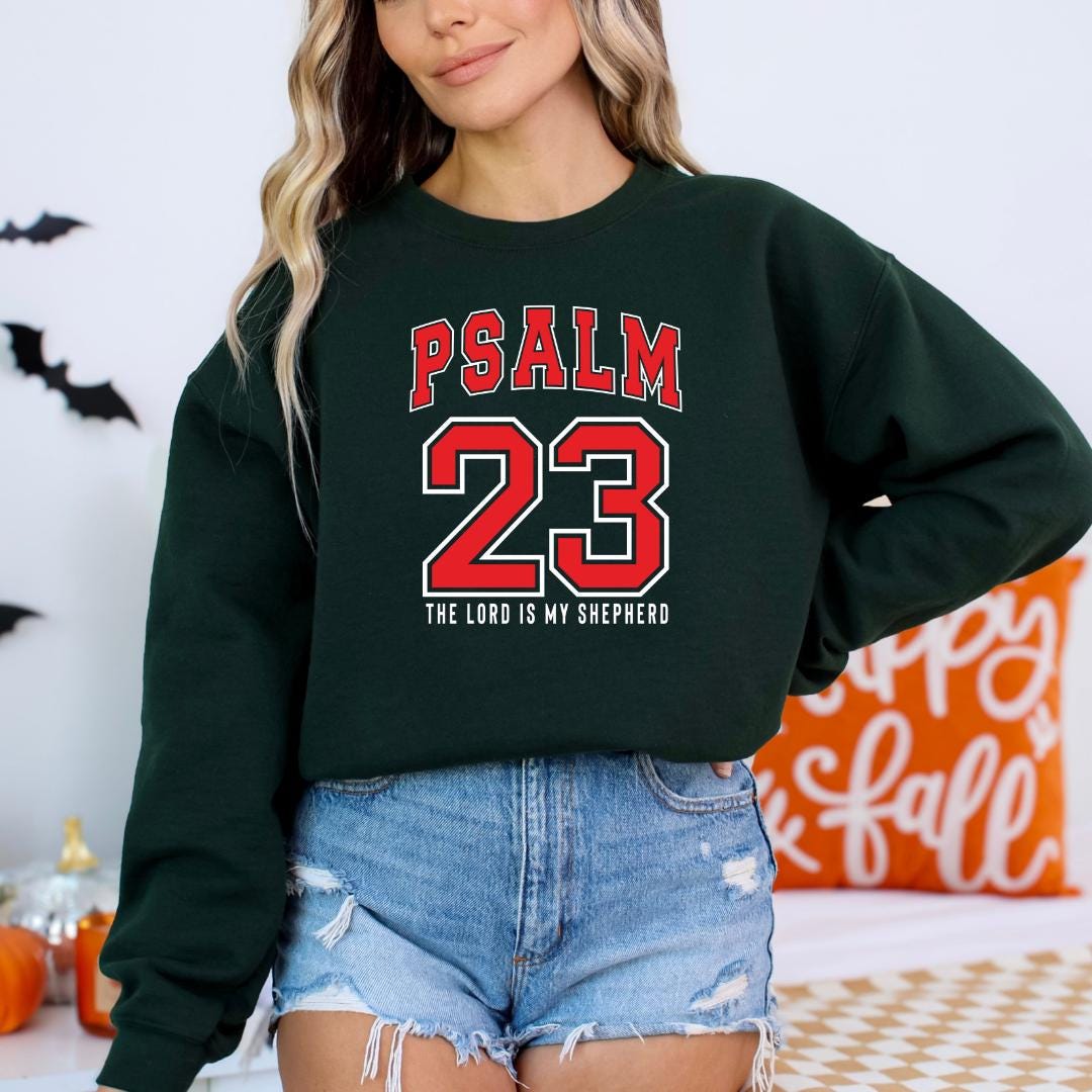 A woman wearing a green sweatshirt with the text "PSALM 23" and "THE LORD IS MY SHEPHERD" printed on it, along with the number 23. She is also wearing denim shorts and a black top.