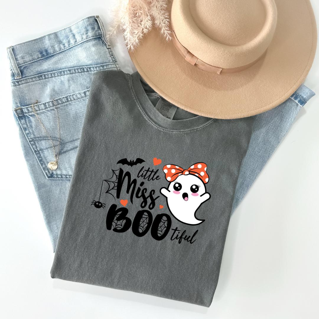 A gray t-shirt with a graphic design featuring a ghost character and the text "Little Miss Boo" printed on it, along with a bow and a heart. The t-shirt is placed on top of a pair of jeans and a beige hat.