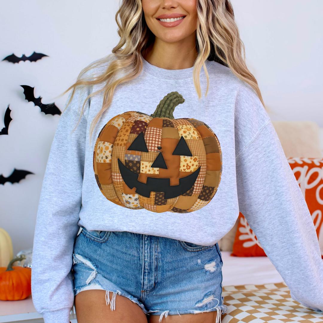 A woman wearing a grey sweatshirt with a patchwork pumpkin design on it, standing in front of a Halloween-themed background.