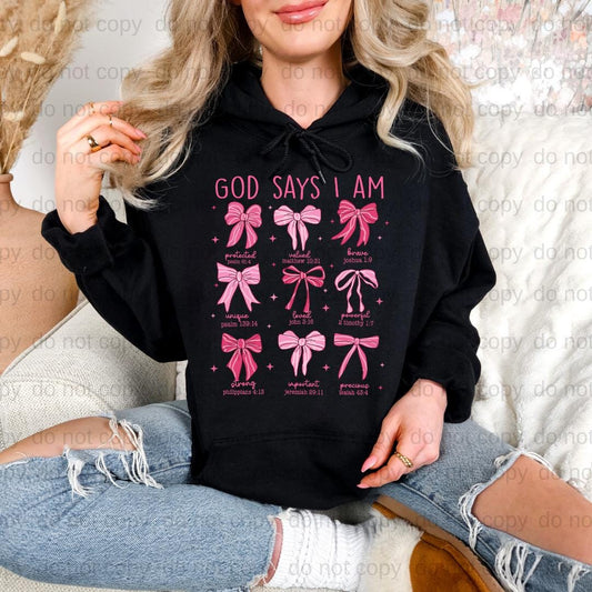 A woman wearing a black hoodie with a graphic design of pink bows and text that says "God says I am".
