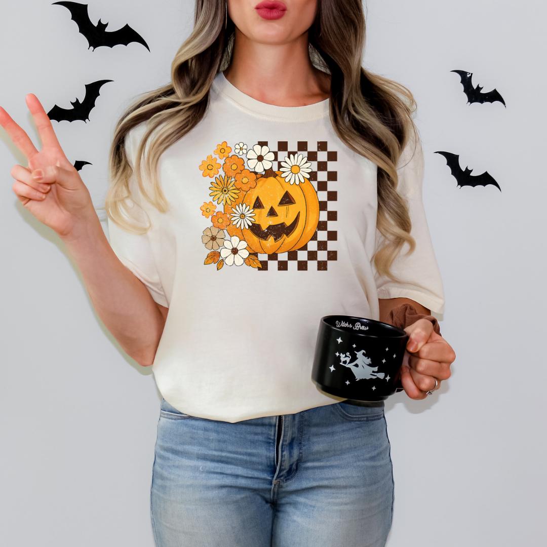 A woman is standing against a white wall, wearing a white t-shirt with a pumpkin and floral design, and holding a black mug with a white design.