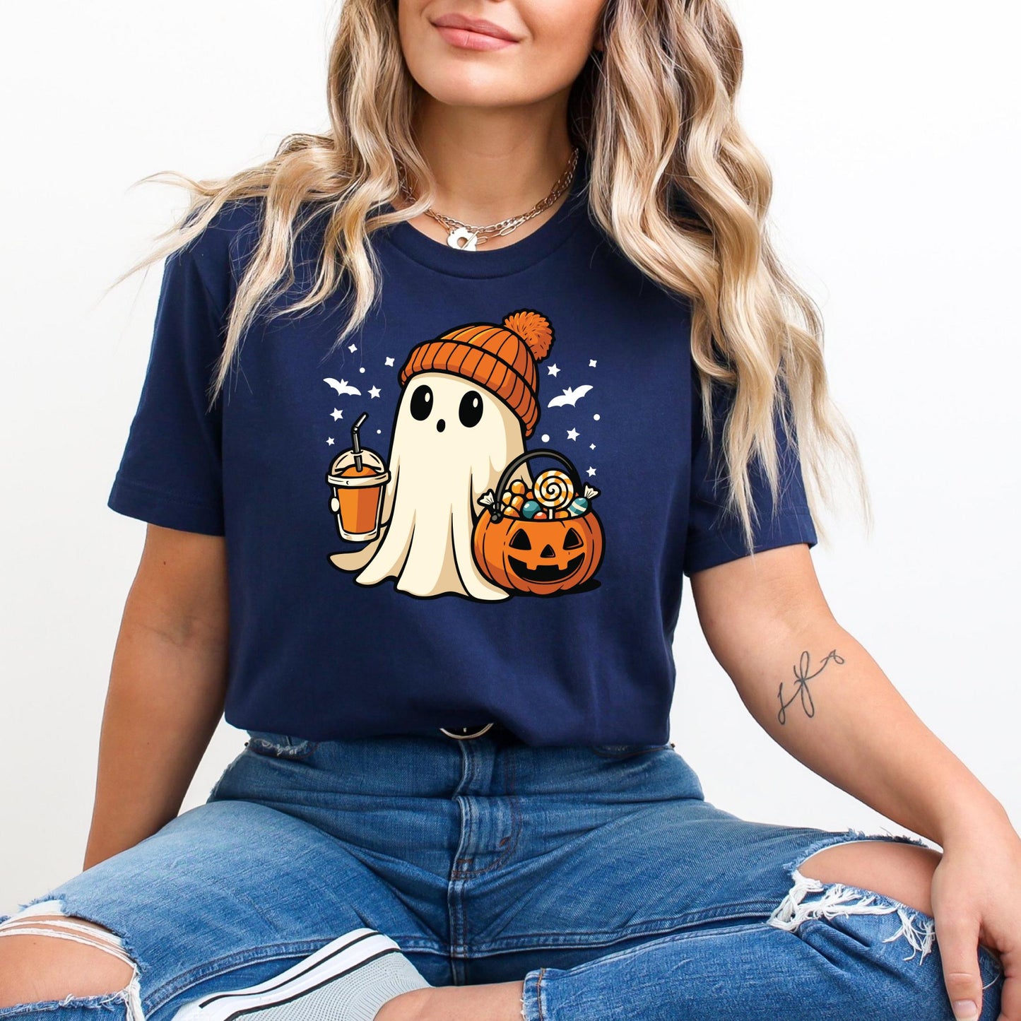 A woman wearing a blue t-shirt with a graphic of a ghost holding a pumpkin and a drink, sitting on the floor with her legs crossed.