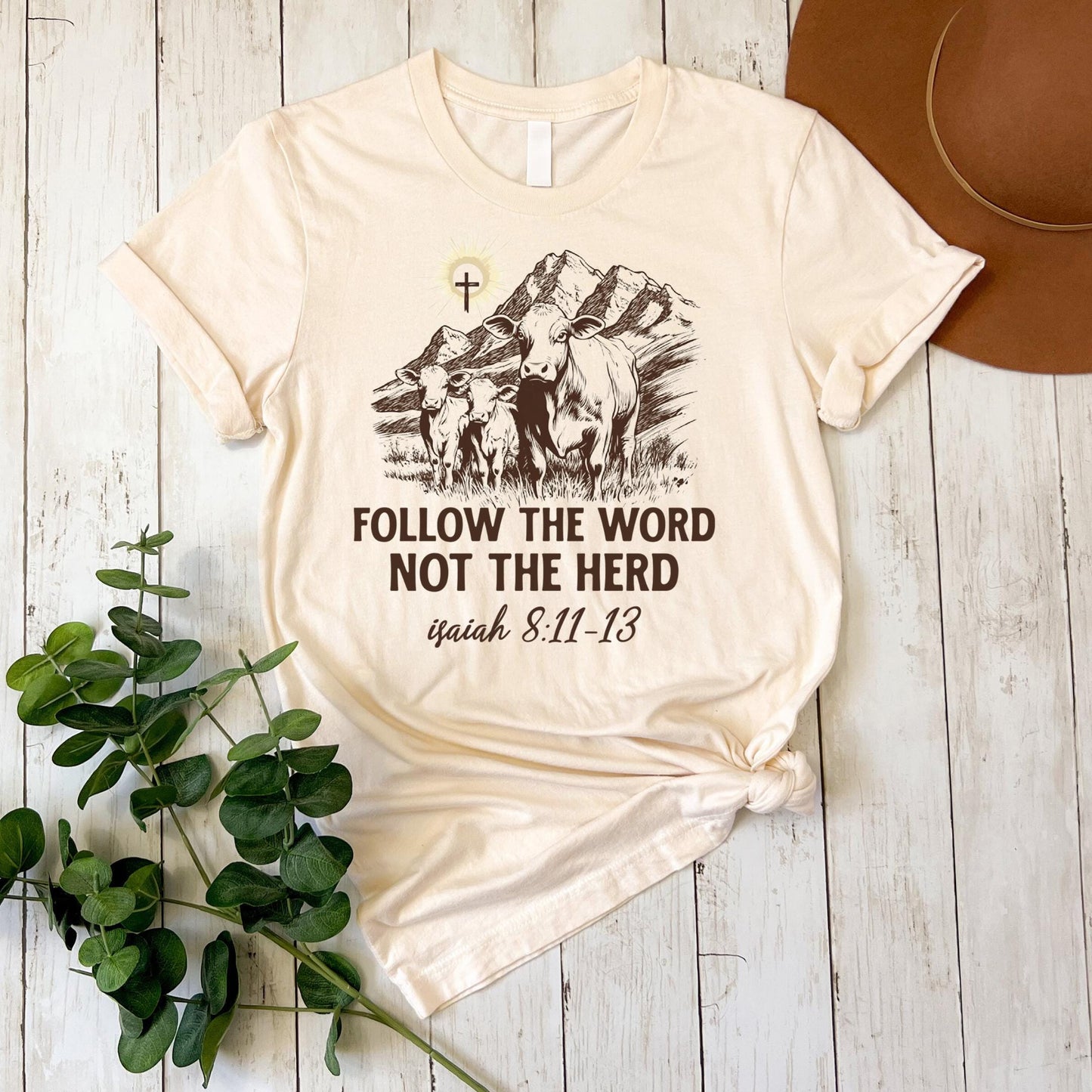 A beige t-shirt with a graphic design and text printed on it, placed on a wooden surface alongside a hat and some greenery.