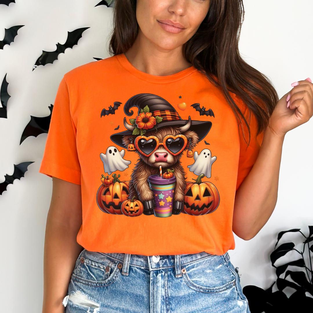 A woman wearing an orange t-shirt with a Halloween-themed graphic of a cow wearing a witch's hat, sunglasses, and holding a cup with a pumpkin and ghost.