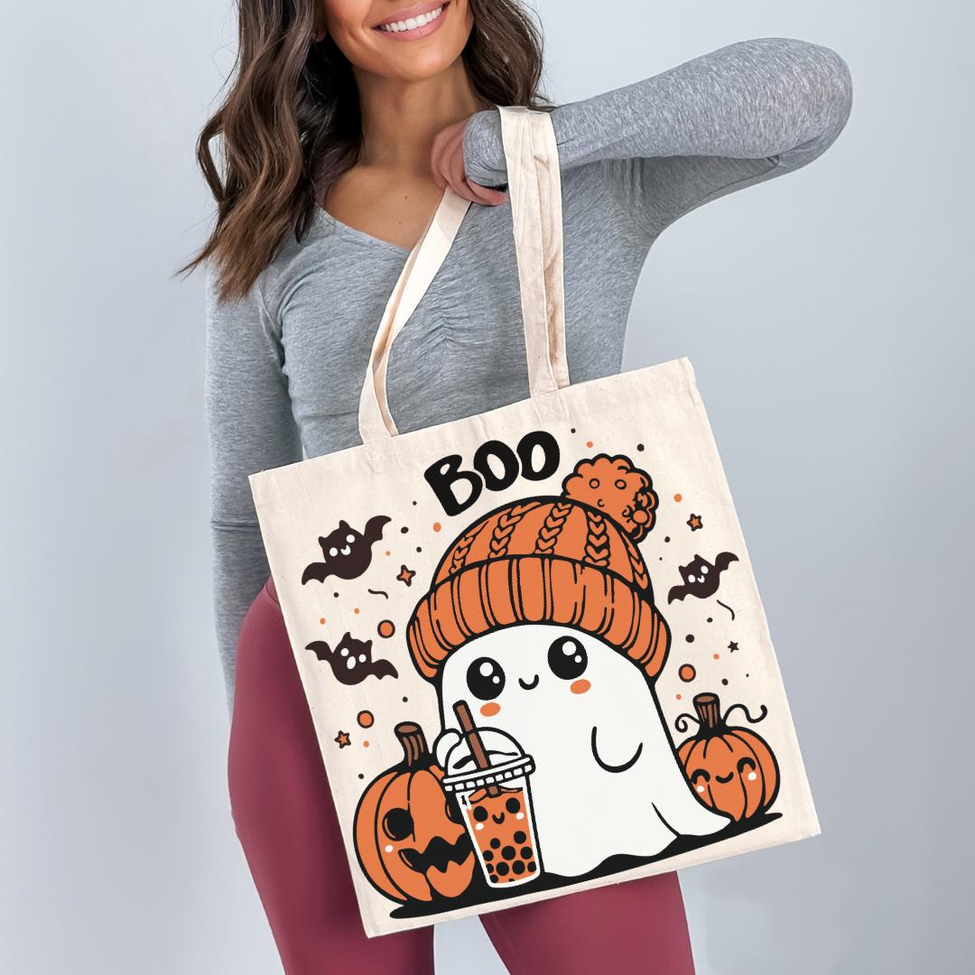 A woman is holding a large tote bag with a Halloween-themed design featuring a ghost, pumpkins, and a drink.