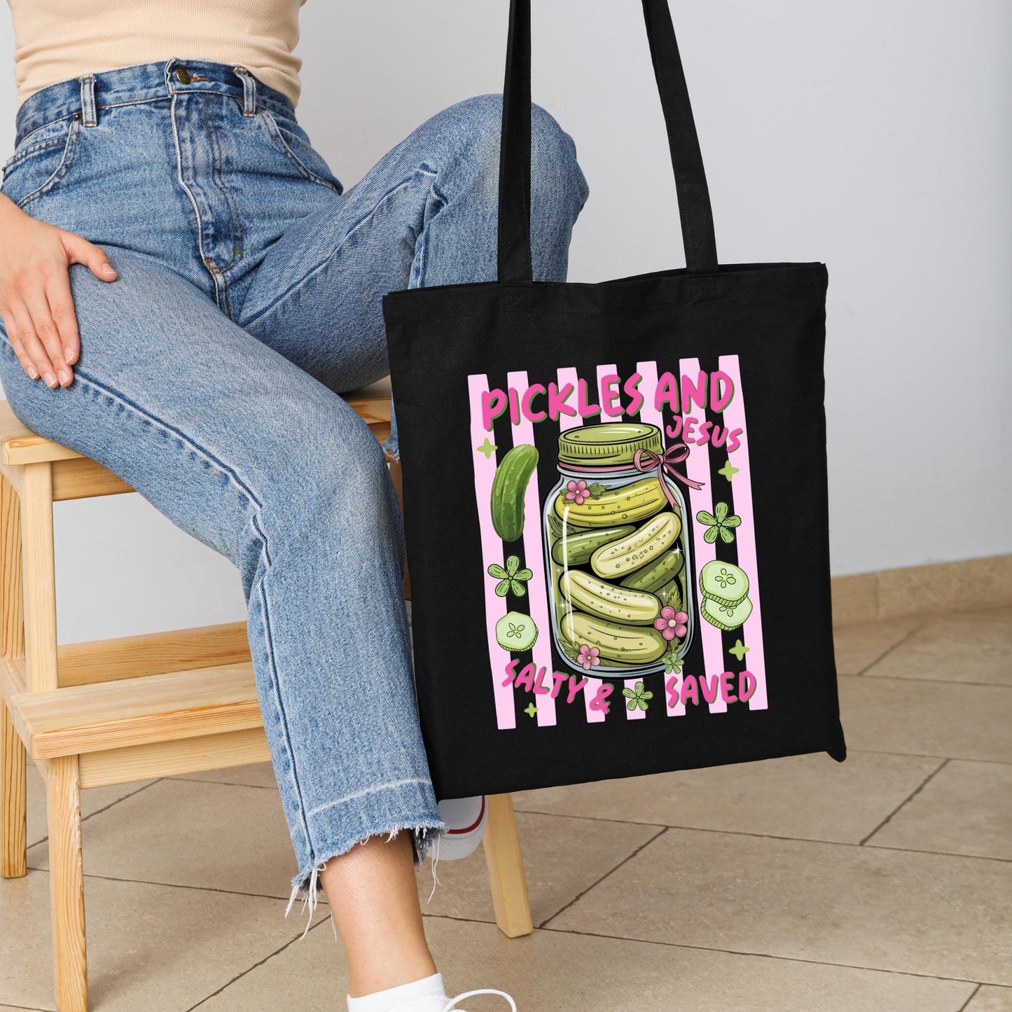 A person is sitting on a wooden chair, holding a black tote bag with a graphic design of a jar of pickles and the text "Pickles and Jesus Salty & Saved".