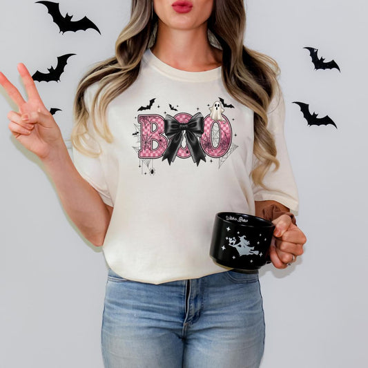 A woman wearing a white t-shirt with the word "BOO" and a bow, holding a black mug with a white design, and surrounded by black bats on a white background.