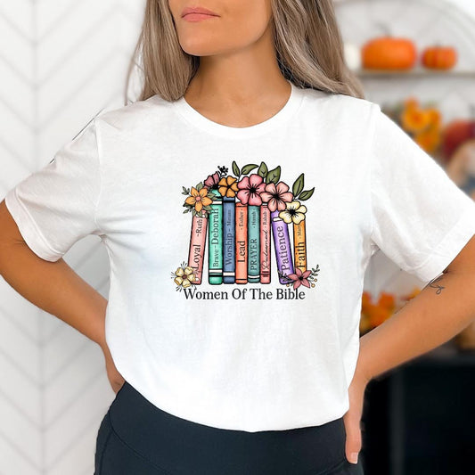 A woman wearing a white t-shirt with a colorful graphic design of a stack of books surrounded by flowers and the text "Women Of The Bible" printed on it.