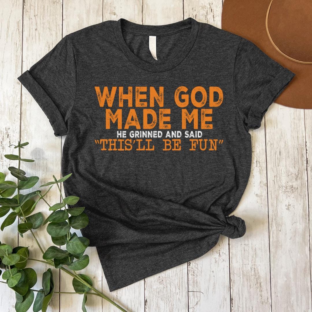A gray t-shirt with orange text that reads "WHEN GOD MADE ME HE GRINNED AND SAID THIS'LL BE FUN'" is displayed on a wooden surface, accompanied by a brown hat and some greenery.