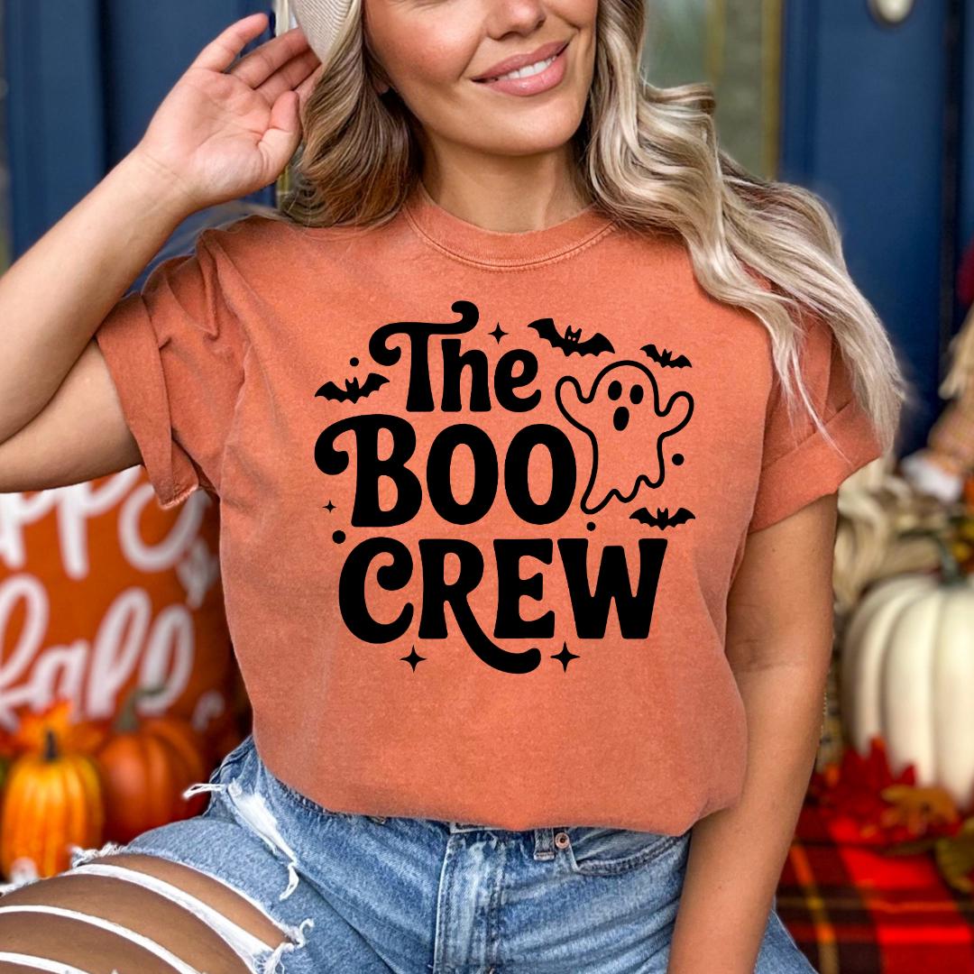 A woman wearing a t-shirt with the text "The Boo Crew" printed on it, smiling and posing in front of Halloween decorations.