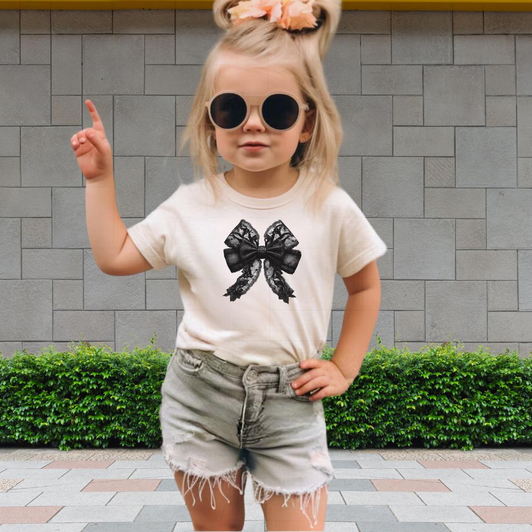 A young girl wearing a white t-shirt with a bow design, sunglasses, and denim shorts is standing in front of a brick wall.