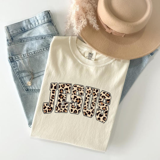 A casual outfit consisting of a white t-shirt with a leopard print design, a pair of blue jeans, and a beige hat is laid out on a white surface.