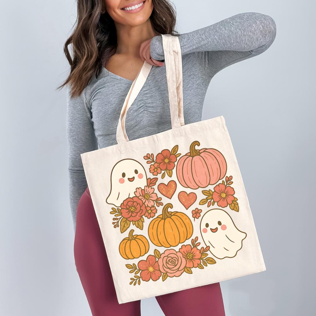 A woman is holding a white tote bag with a Halloween-themed design featuring ghostly figures, pumpkins, flowers, and hearts.