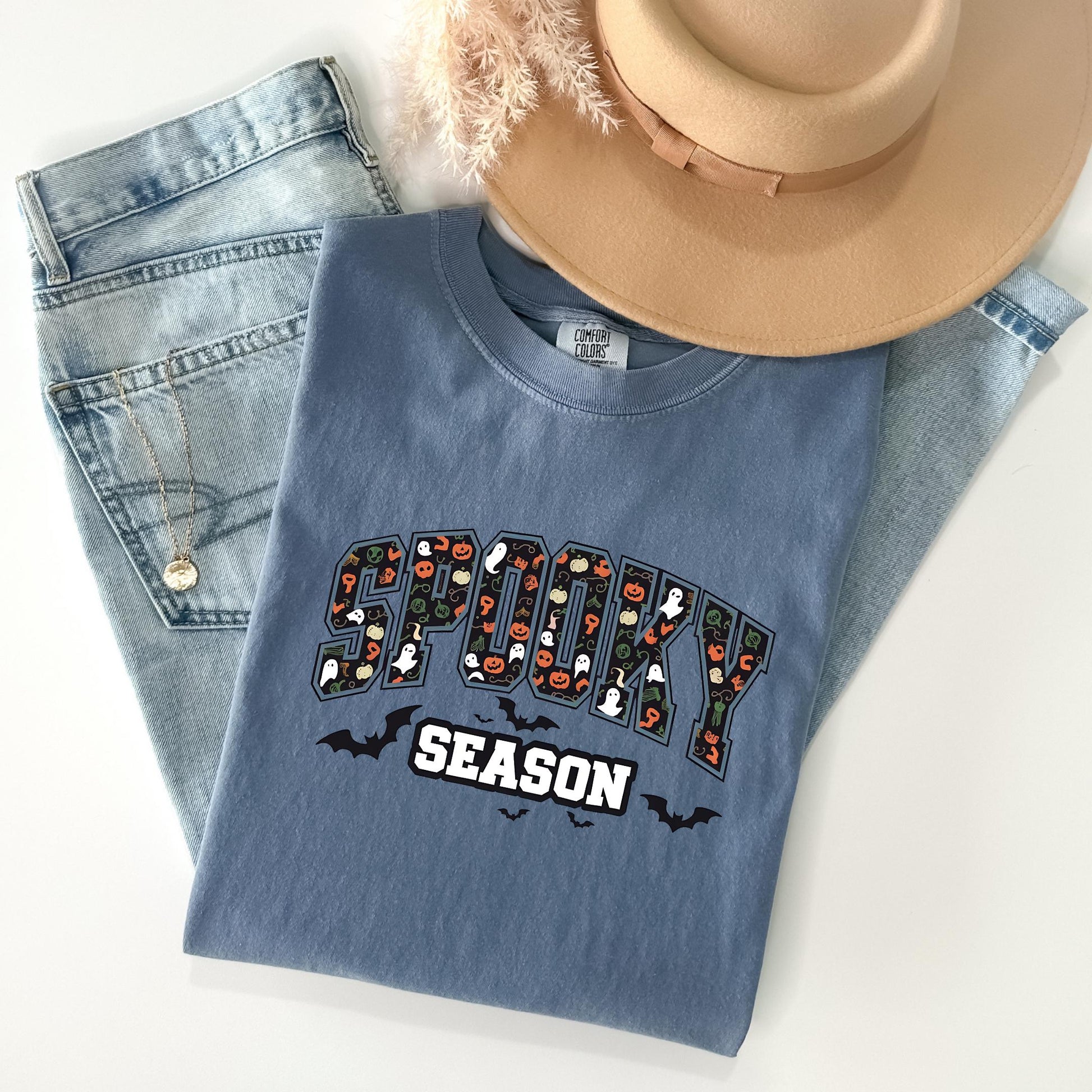 A blue t-shirt with the word "SPOOKY" printed on it, along with Halloween-themed graphics, is laid out on a white surface. The t-shirt is accompanied by a pair of jeans and a beige hat.