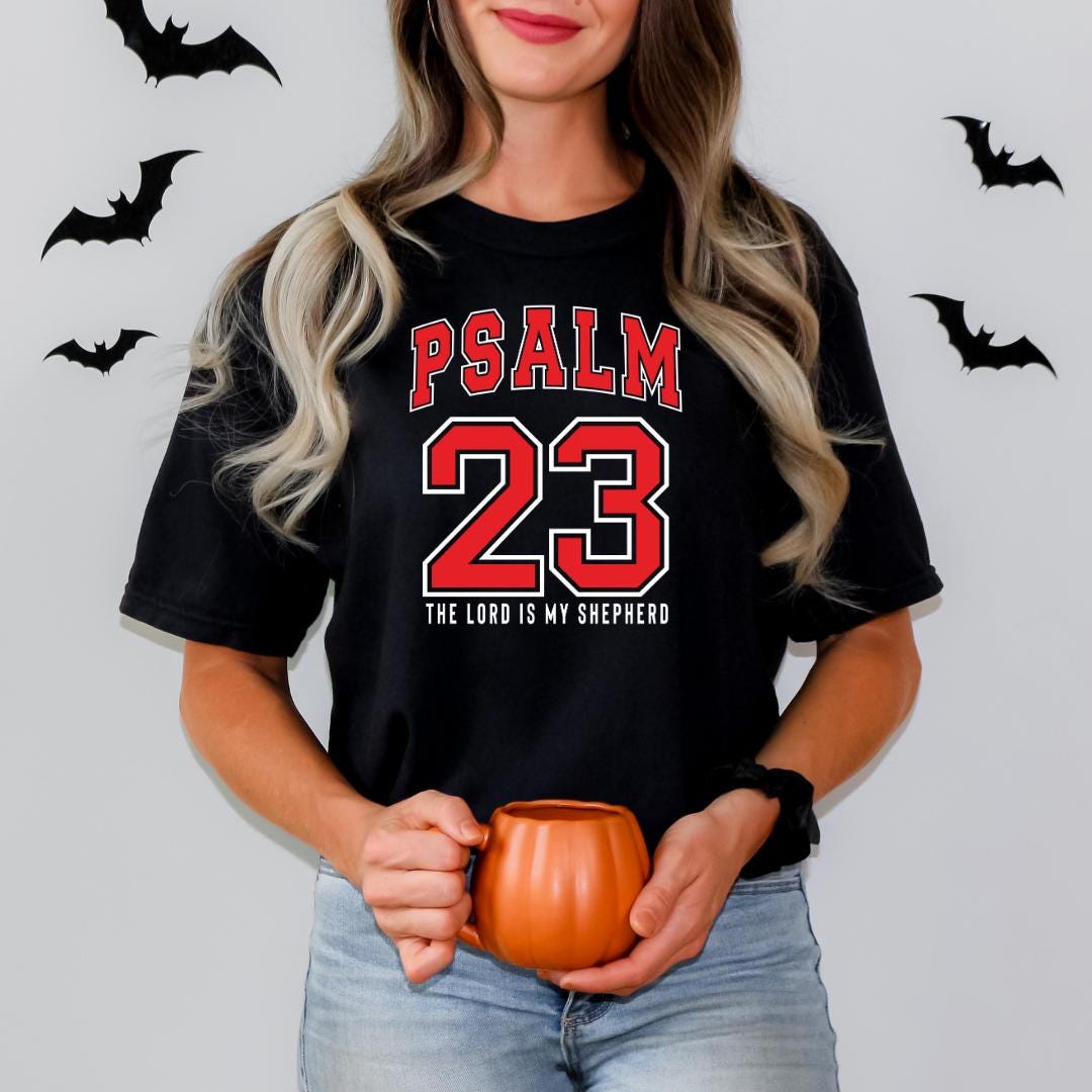 A woman with long blonde hair is holding a pumpkin and a mug, wearing a black t-shirt with the text "PSALM 23" and "THE LORD IS MY SHEPHERD" printed on it.