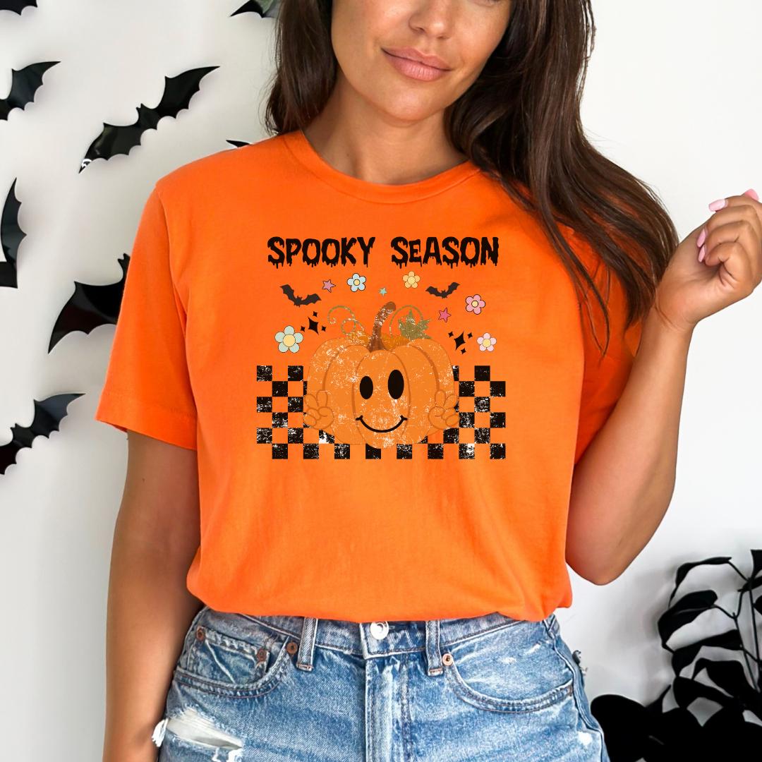 A woman wearing an orange t-shirt with a pumpkin graphic and the text "Spooky Season" printed on it, standing in front of a wall with black bats.