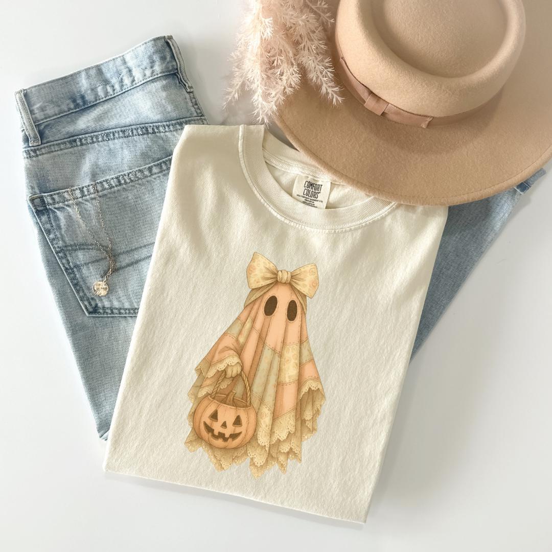 A white t-shirt with a Halloween-themed design, a pair of blue jeans, and a beige hat are arranged on a white surface.