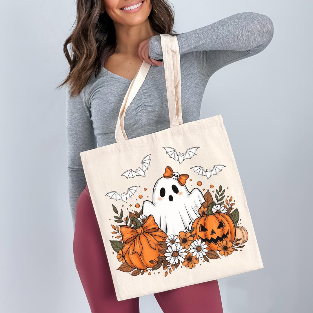 A woman is holding a white tote bag with a Halloween-themed design featuring a ghost, pumpkins, flowers, and bats.