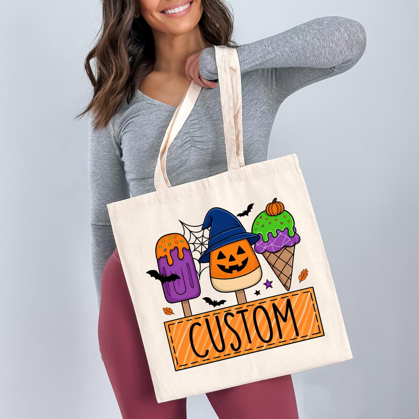 A woman is holding a white tote bag with a Halloween-themed design featuring three colorful popsicles and a witch hat.