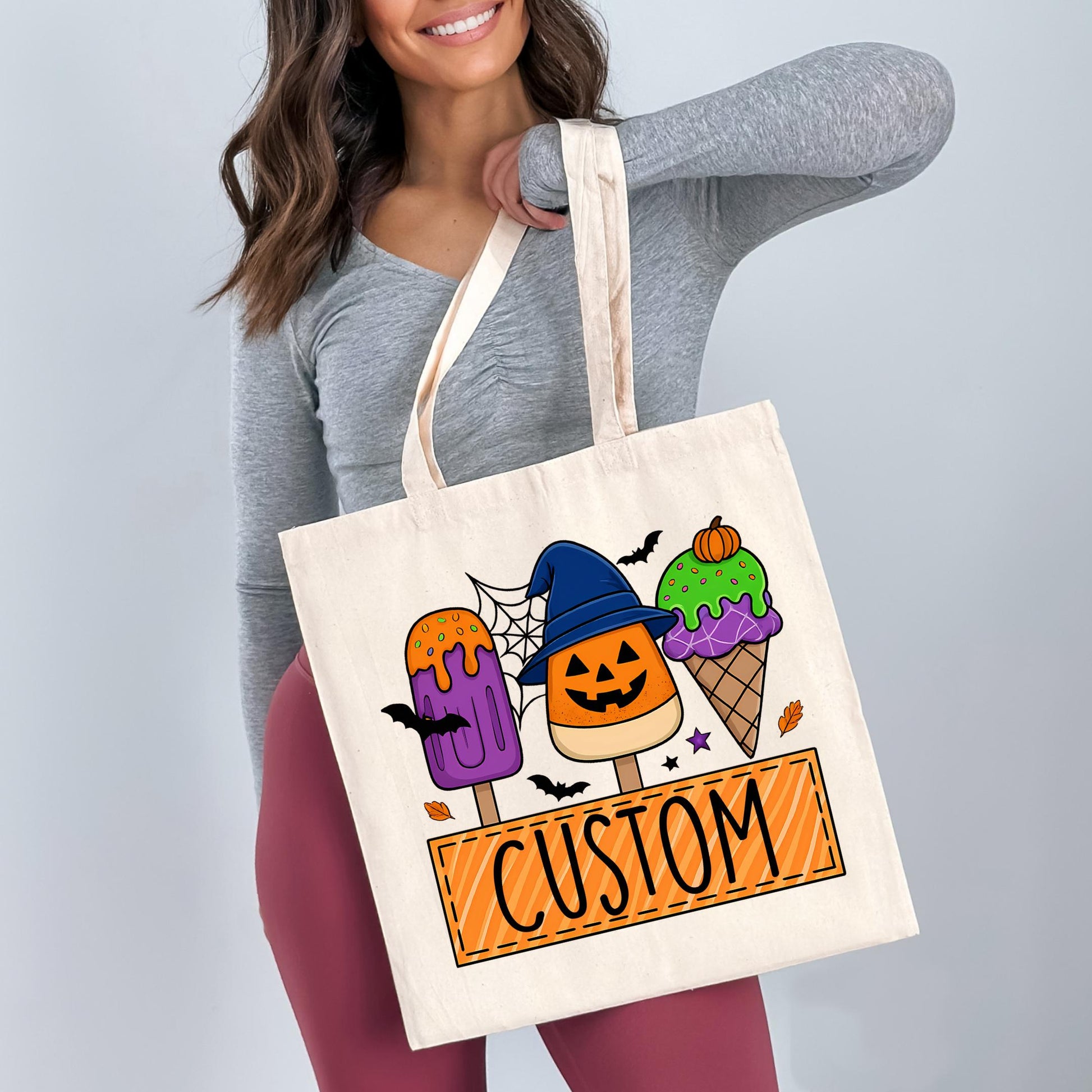 A woman is holding a white tote bag with a Halloween-themed design featuring three colorful popsicles and a witch hat.