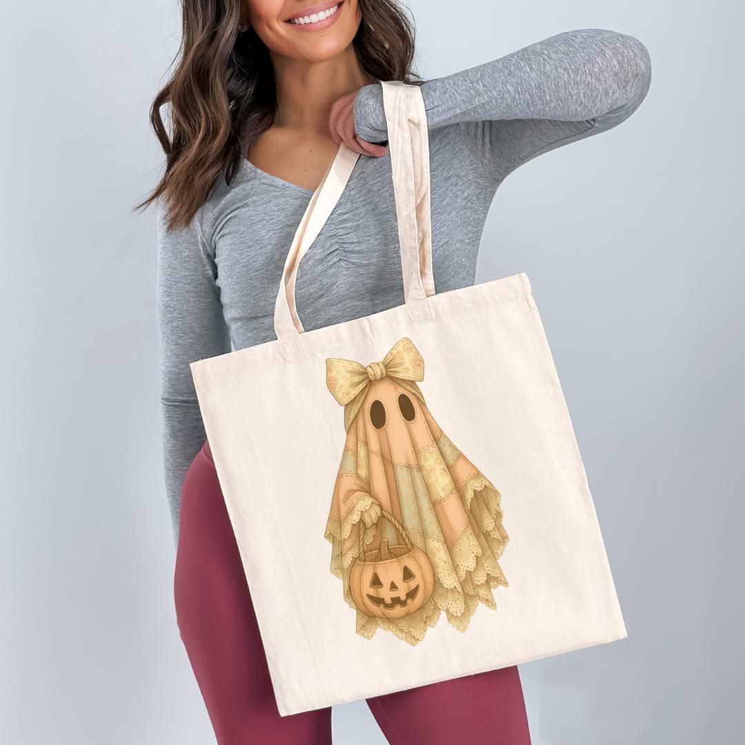 A woman is holding a white tote bag with a Halloween-themed design featuring a ghost, a pumpkin, and a bow.