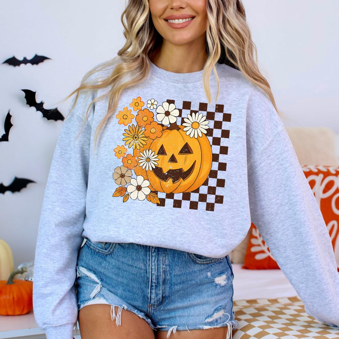 A woman wearing a grey sweatshirt with a pumpkin and flower design on it, standing in front of a Halloween-themed background.