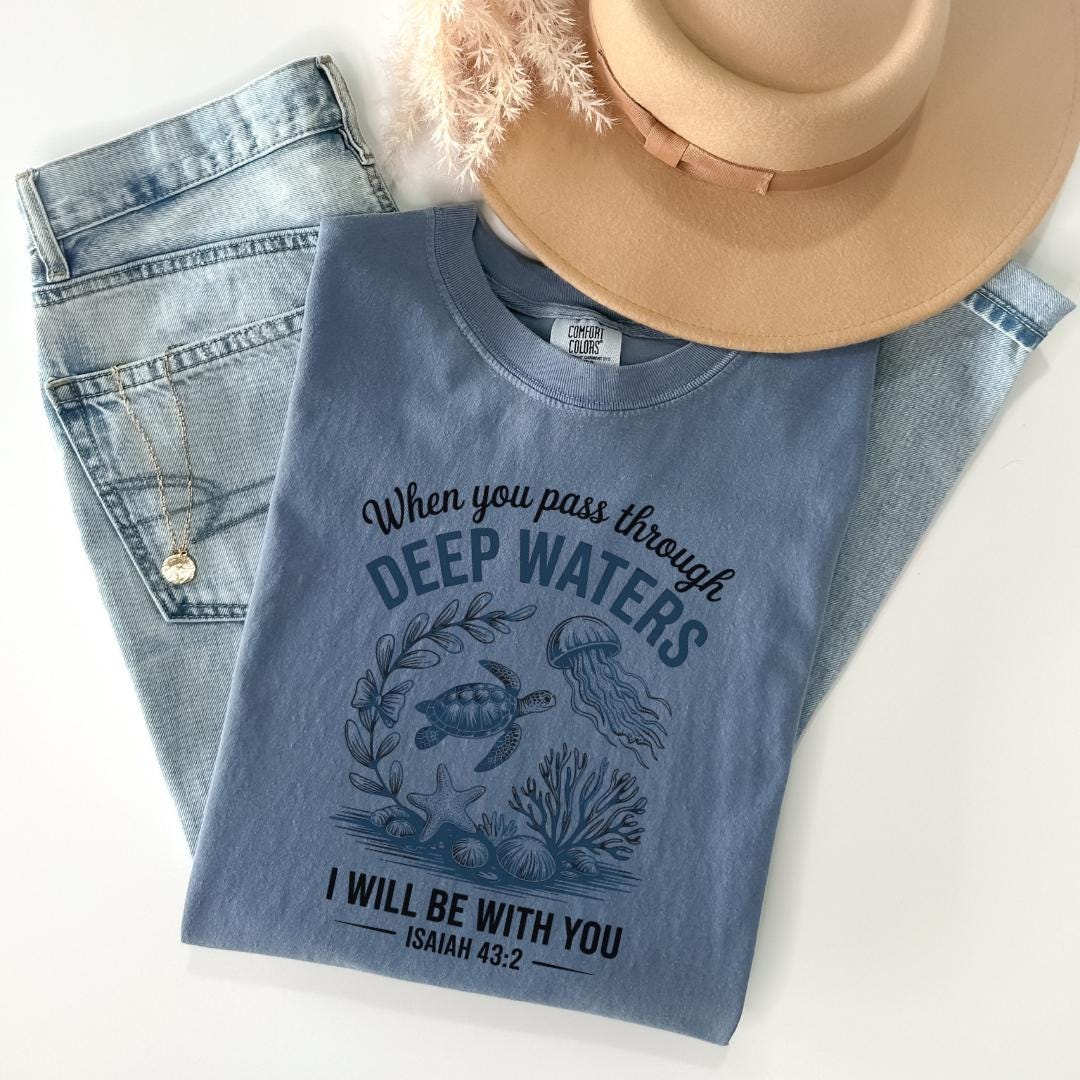 A blue t-shirt with a graphic design of marine life, including a turtle, jellyfish, and seaweed, along with the text "When you pass through DEEP WATERS I WILL BE WITH YOU ISAIAH 43:2". The t-shirt is placed on top of a pair of jeans and a beige hat, and there is a necklace visible in the background.