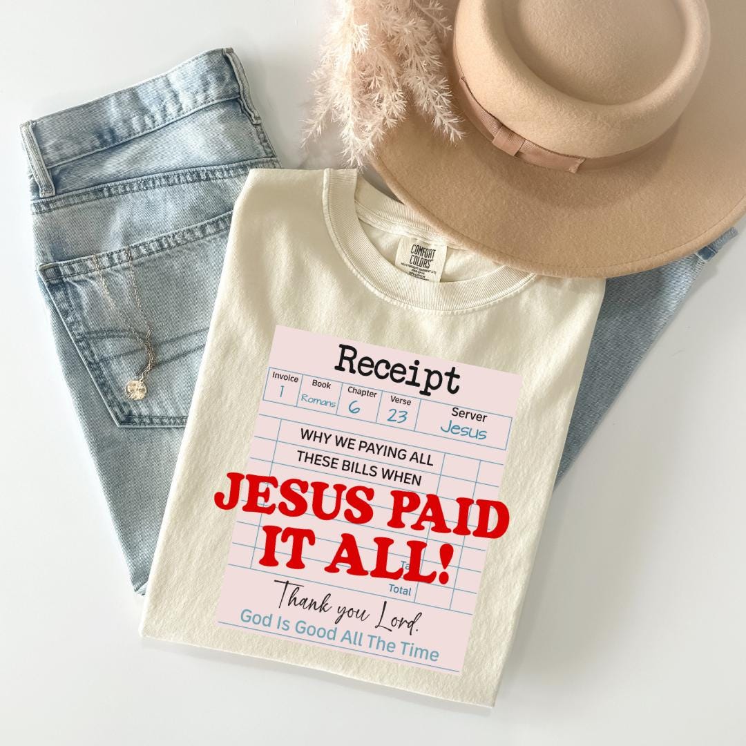 A white t-shirt with a humorous text printed on it, along with a pair of jeans and a beige hat, all laid out on a white surface.