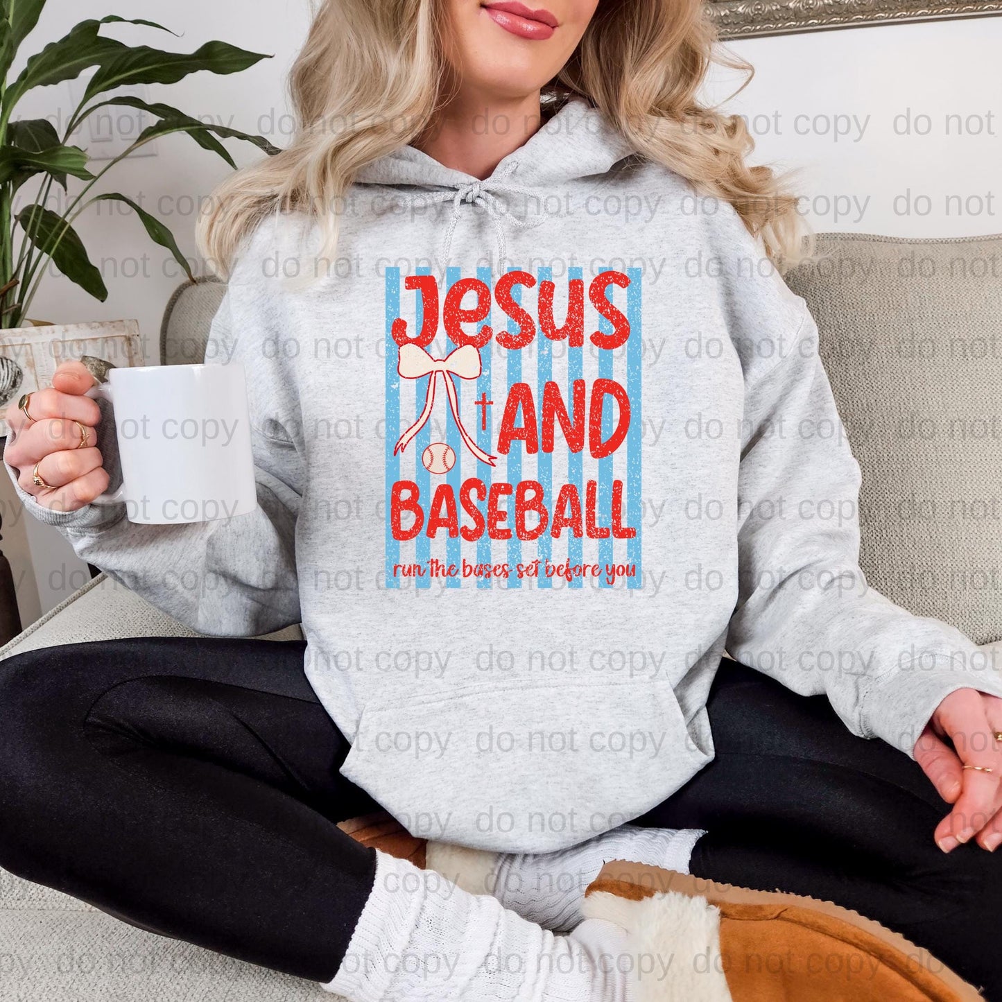 A woman sitting on a couch wearing a gray hoodie with a graphic design that says "Jesus and Baseball run the bases & set before you".