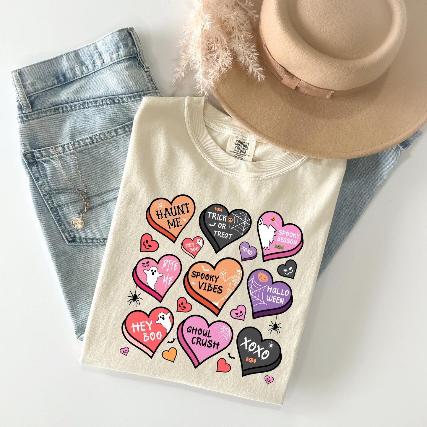 a white t-shirt with a variety of Halloween-themed hearts and text, including phrases like "HAINT ME", "TRICK OR TRUST", "SPOOKY VIBES", "HELL O'LUCKY", "HEY BOO", "SHOUL CRUSH", "XO", and "HEY BOO". The t-shirt is laid out on a white surface, with a pair of jeans and a beige hat visible in the background.