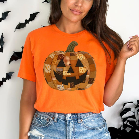 A woman wearing an orange t-shirt with a patchwork pumpkin design on it, standing in front of a wall with black bats.