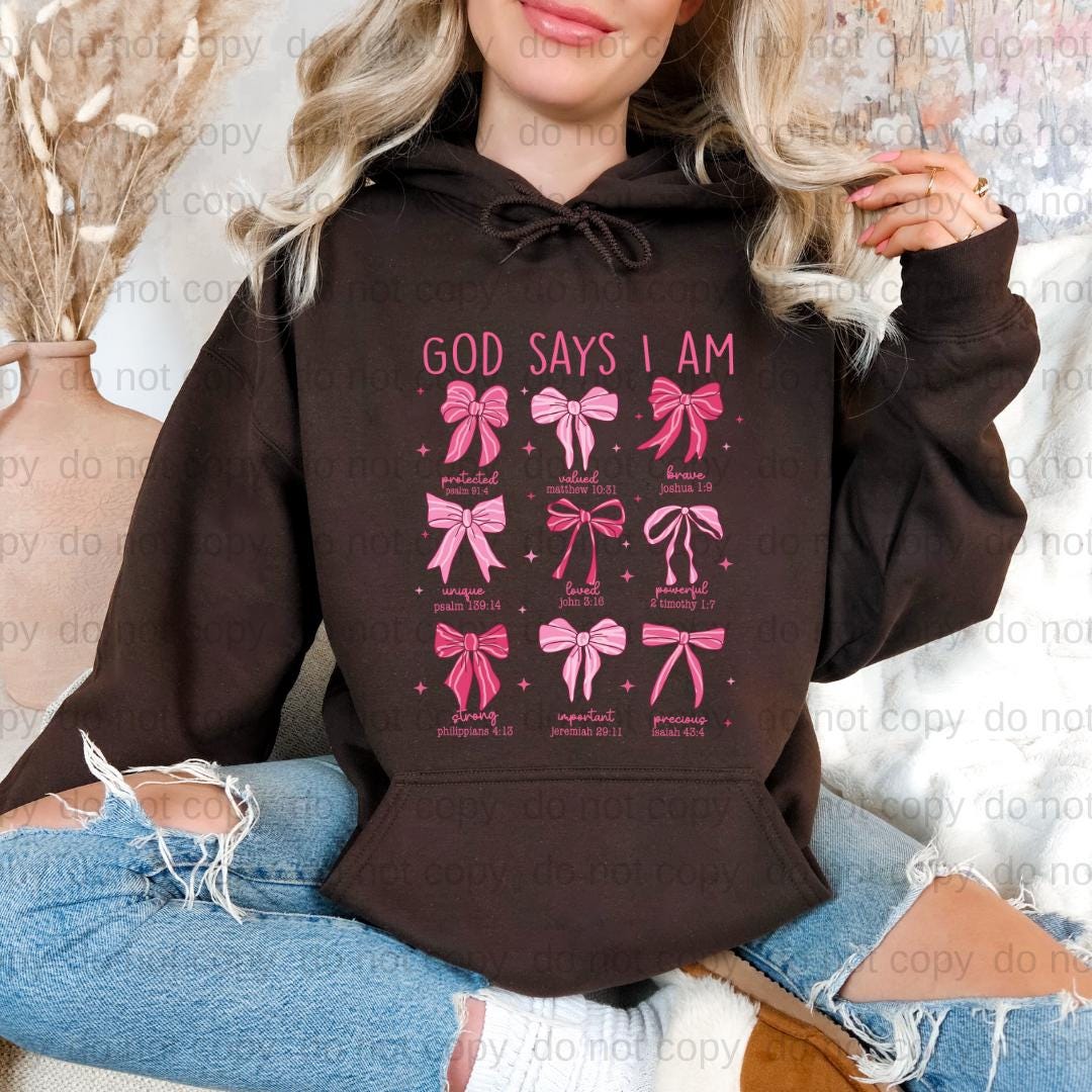 A woman wearing a black hoodie with a graphic design of pink bows and text that says "GOD SAYS I AM".