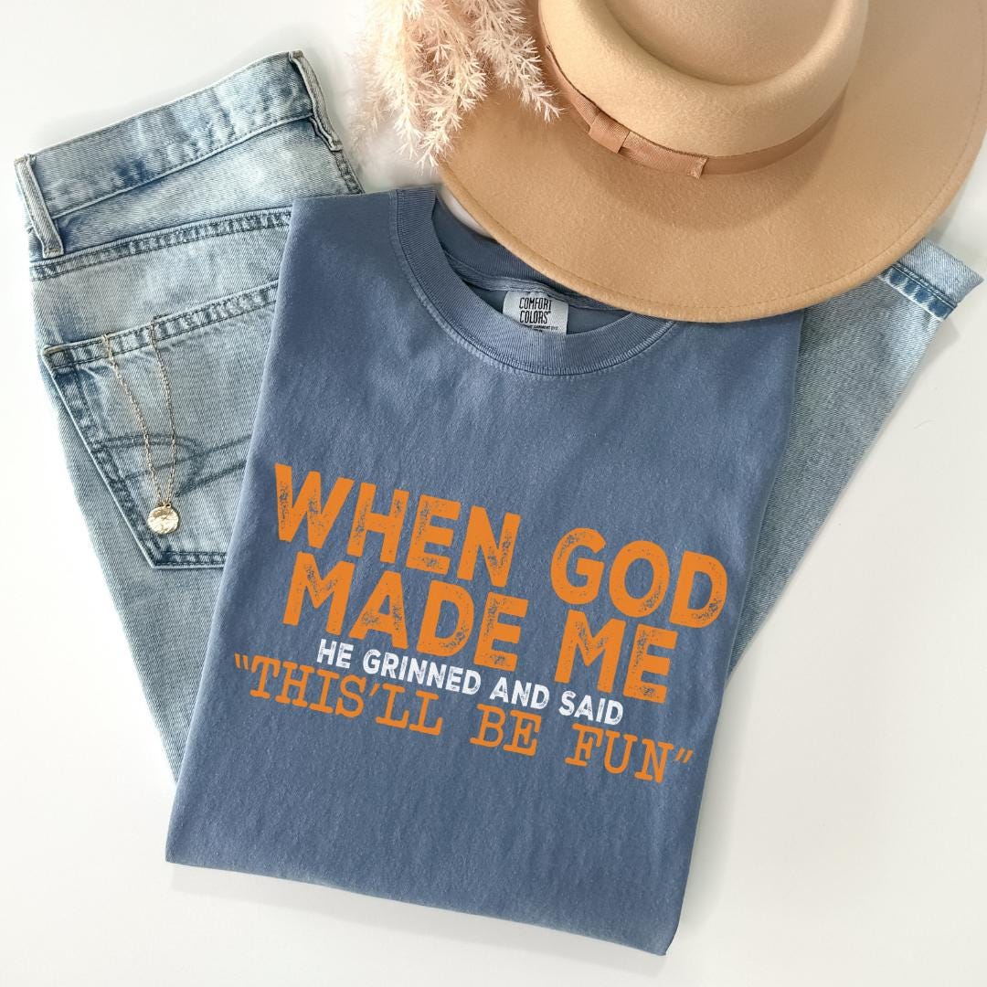 A blue t-shirt with orange text, a pair of jeans, and a beige hat are arranged on a white background.