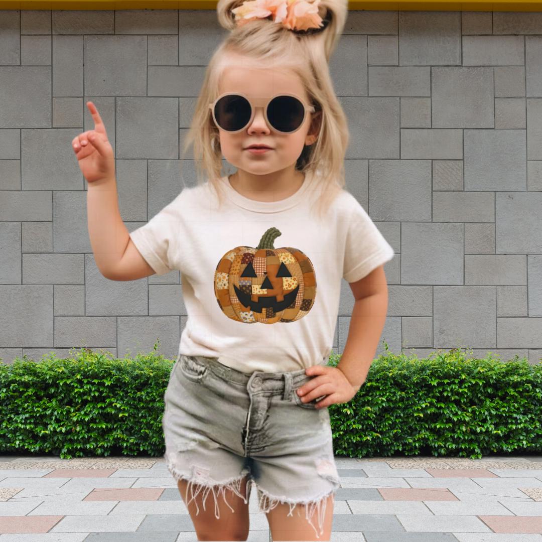 A young girl wearing a white t-shirt with a pumpkin graphic, sunglasses, and denim shorts is standing in front of a brick wall.