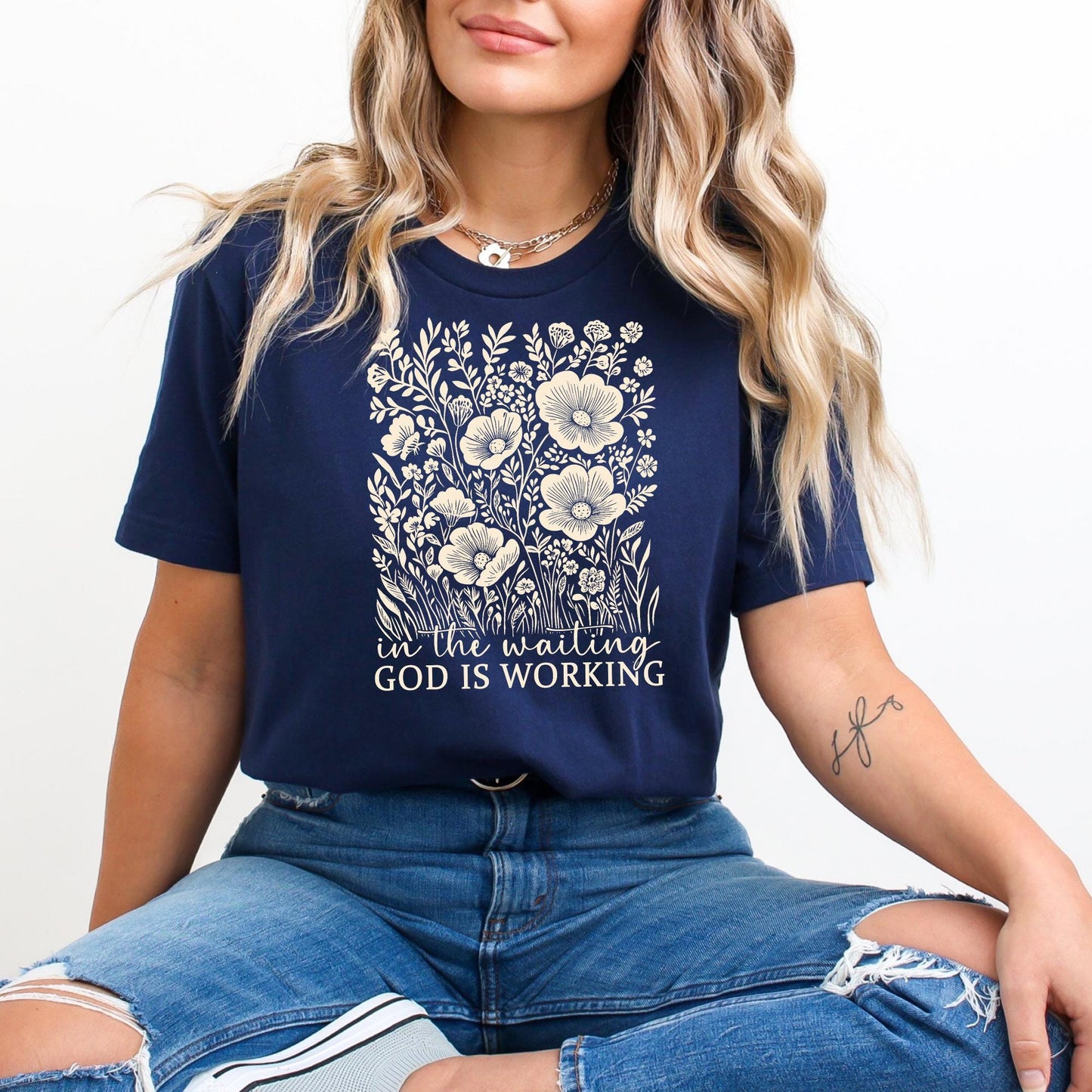 A woman wearing a navy blue t-shirt with a floral design and the text "God is working" is sitting on the floor.