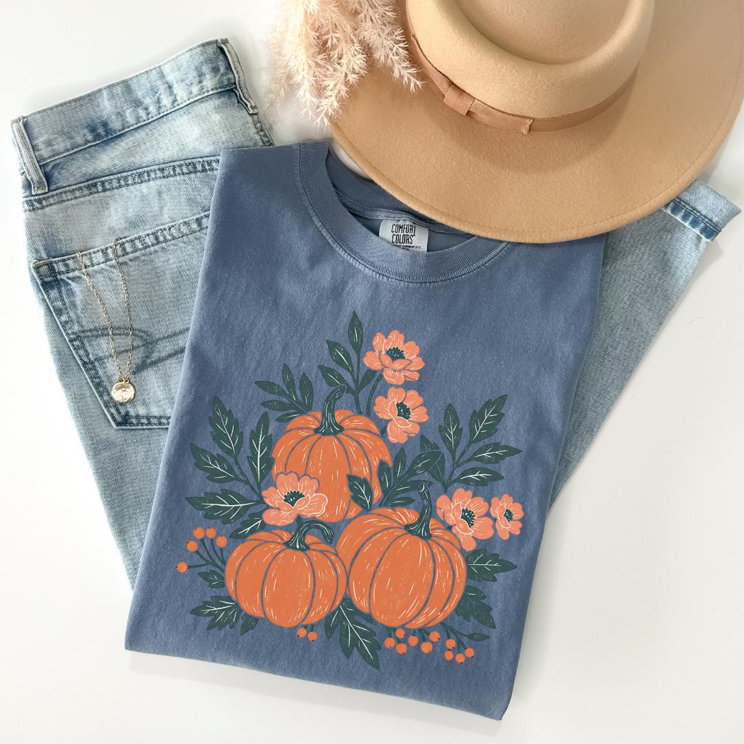 A casual outfit consisting of a blue t-shirt with a pumpkin and leaf design, a pair of blue jeans, and a beige hat is laid out on a white surface.