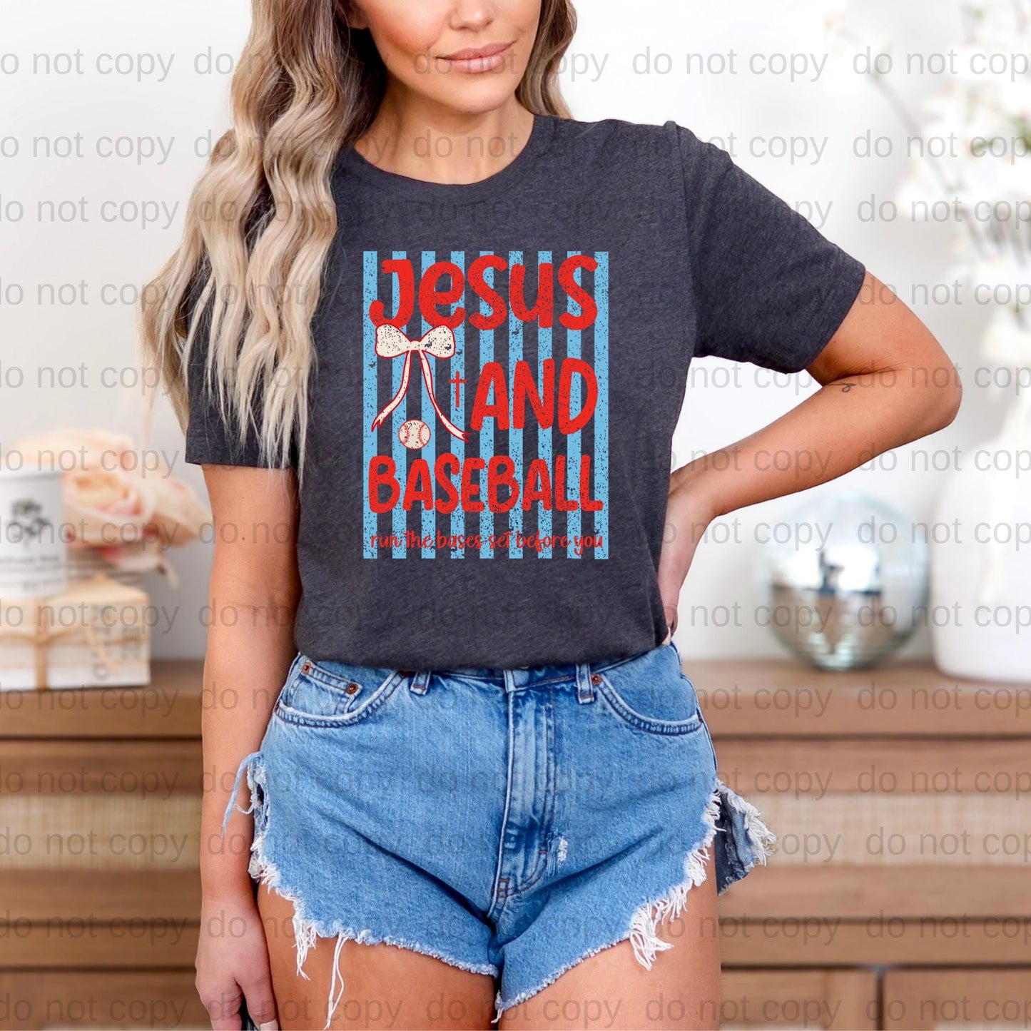 A woman wearing a gray t-shirt with a graphic design and denim shorts stands in front of a wooden dresser.
