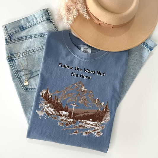 A blue t-shirt with a mountain landscape design and the text "Follow the Word Not the Herd" printed on it, along with a beige hat and a pair of blue jeans.
