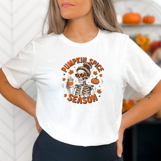 A woman wearing a white t-shirt with a graphic design featuring a skeleton wearing sunglasses and holding a pumpkin, surrounded by autumn leaves and the text "Pumpkin Spice Season".