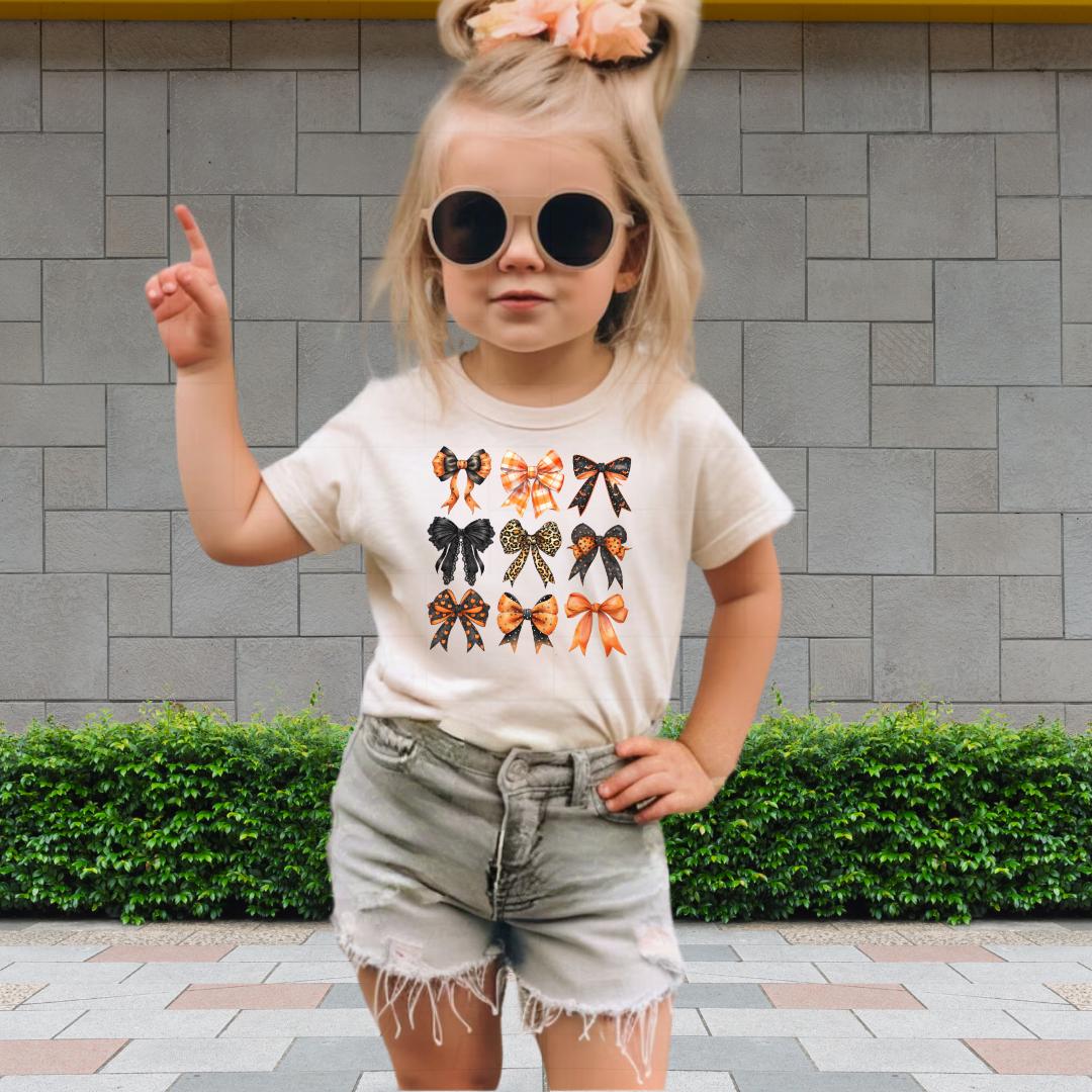 A young girl wearing a white t-shirt with bow designs, sunglasses, and denim shorts is standing in front of a brick wall.