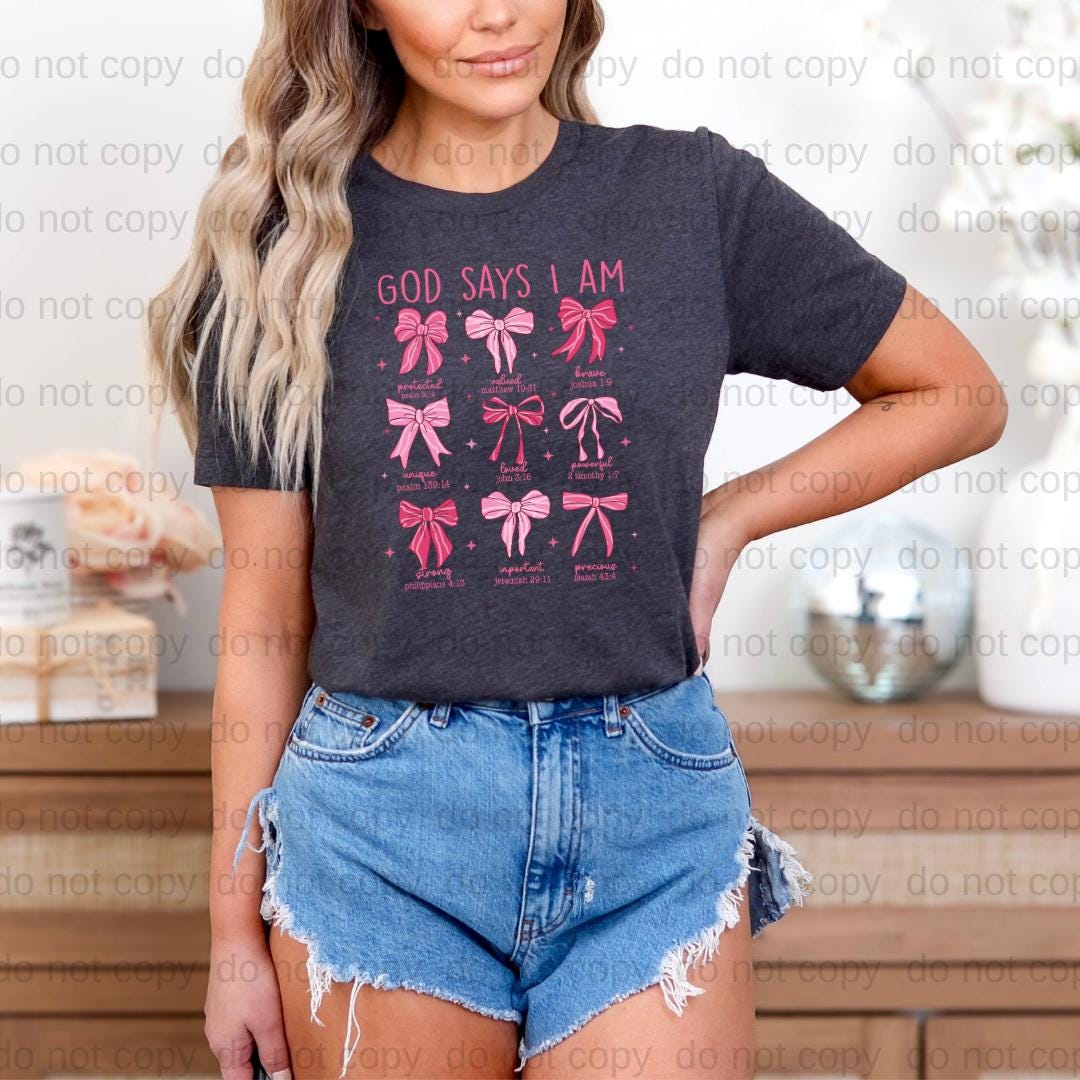 A woman wearing a black t-shirt with a graphic design of pink bows and the text "GOD SAYS I AM" printed on it. She is also wearing denim shorts.