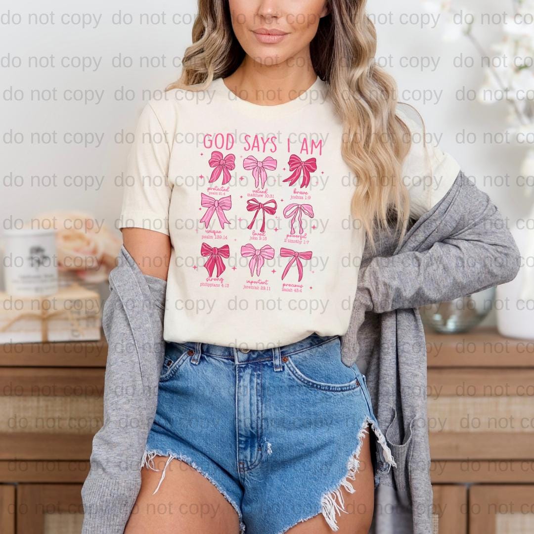 A woman wearing a white t-shirt with a graphic design of pink bows and the text "GOD SAYS I AM" printed on it, paired with denim shorts and a gray cardigan.